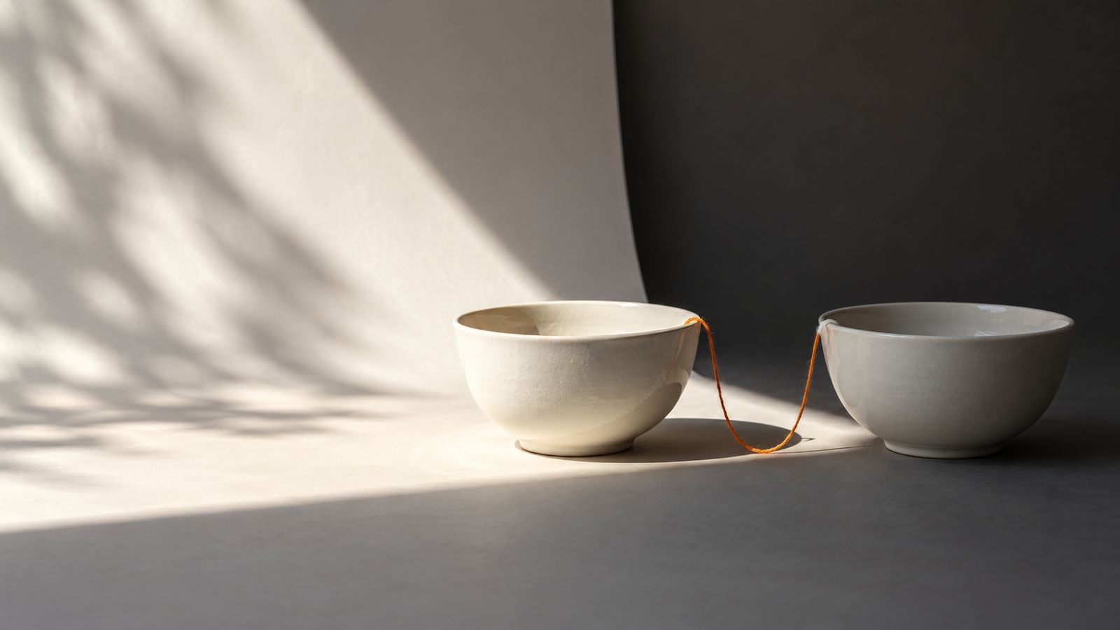 Two white ceramic bowls joined by a thin orange thread on a grey surface