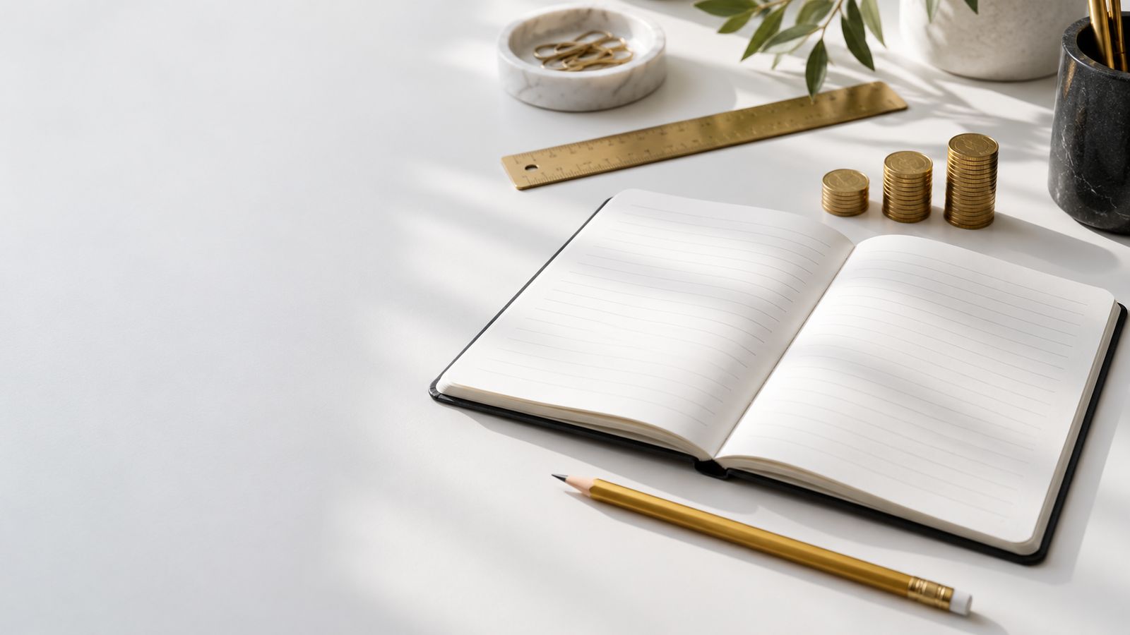 An open notebook, a pencil, a brass ruler and small stacks of coins on a white desk